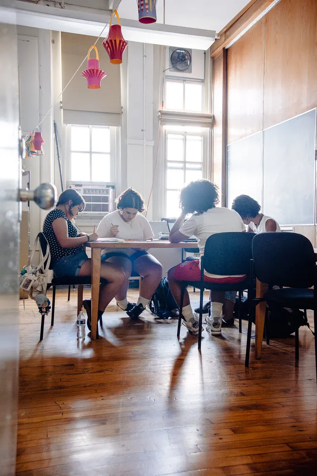 Four students working quietly around a table in a sunlit classroom with tall windows, high ceilings and paper lanterns strung overhead